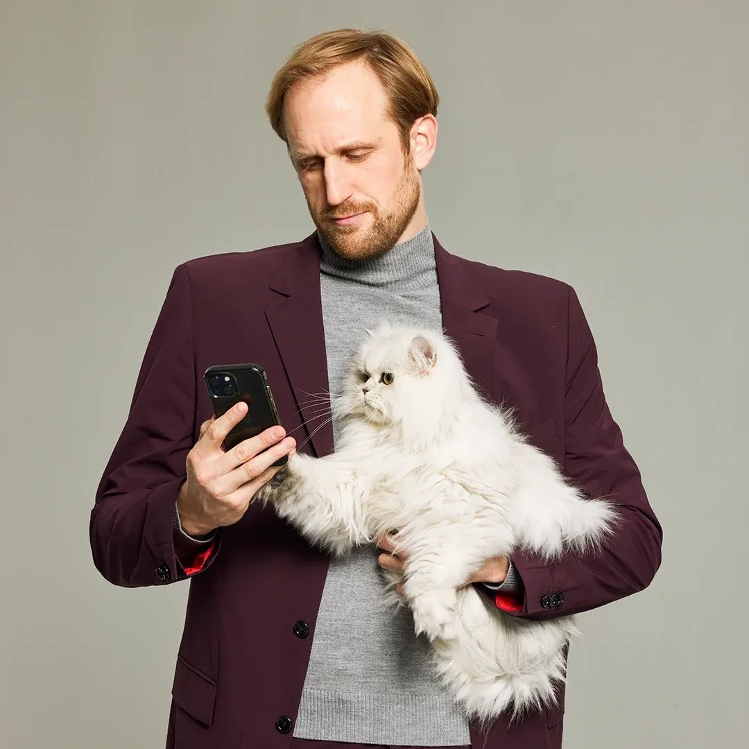 Ein Mann in einem kastanienbraunen Blazer hält eine flauschige weiße Katze, während er auf sein Smartphone schaut. Die Katze greift nach dem Telefon.
