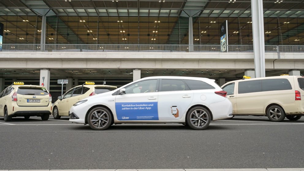 Taxen warten vor dem Flughafen BER, ein Uber-Fahrzeug fährt vorbei