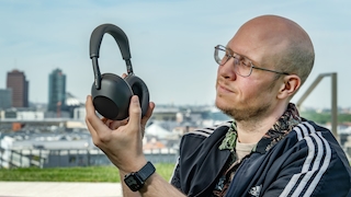 Ein Mann hält die schwarzen Sony WH-1000XM6 Kopfhörer vor seinem Gesicht und betrachtet sie interessiert. Im Hintergrund ist eine unscharfe Silhouette der Stadt Berlin mit mehreren hohen Gebäuden zu erkennen.