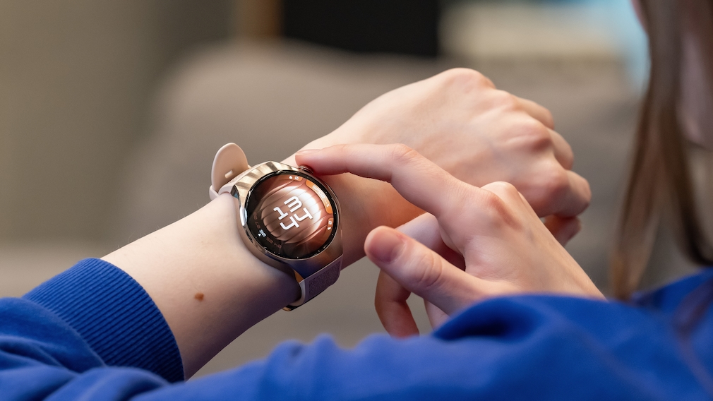 Eine Person trägt eine runde Smartwatch mit roségoldenem Gehäuse und hellrosafarbenem Armband am linken Handgelenk. Auf dem Display wird die Uhrzeit in weißer Digitalanzeige vor einem rötlich-schimmernden Hintergrund dargestellt. Die Person tippt mit dem rechten Zeigefinger auf das Display. Im Hintergrund sind unscharfe Möbelstücke und eine gemütliche Innenraumatmosphäre zu erkennen.