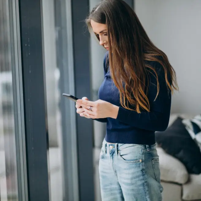 Eine Frau steht traurig am Fenster und schaut auf ihr Handy.