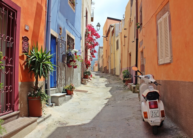 Eine Gasse in Italien, auf beiden Seiten bunte Häuserfassaden mit Blumenschmuck, rechts parkt ein weißer Roller.